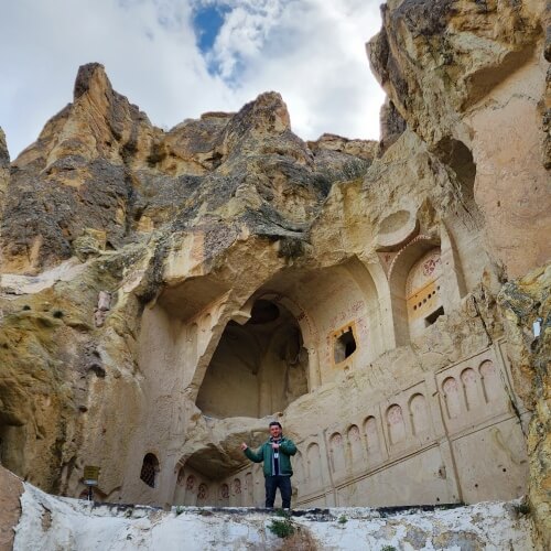Göreme Açık Hava Müzesi’nde yer alan Karanlık Kilise’nin dış cephesi, kaya oyma mimarisi ve detaylı işlemeleriyle birlikte bir kişinin önünde poz verdiği şekilde görülüyor.